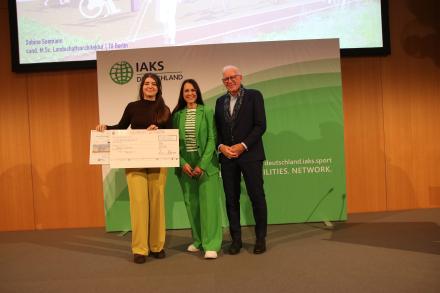 3 Menschen auf Bühne mit Urkunde und Scheck