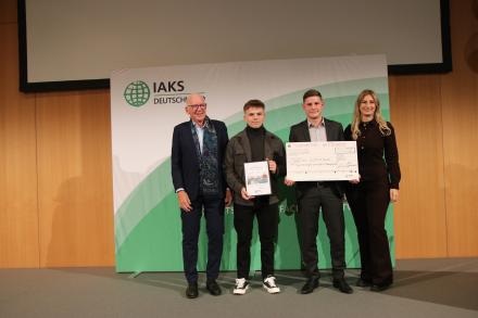 3 Menschen auf Bühne mit Urkunde und Scheck