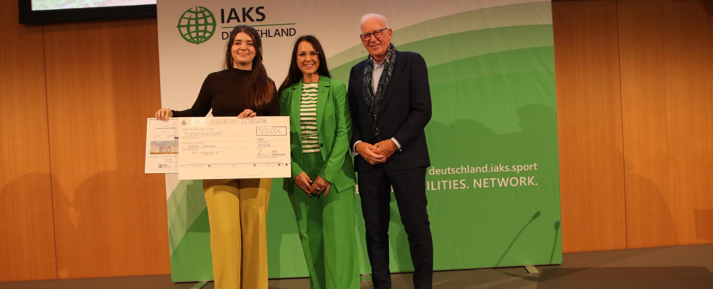 3 Menschen auf Bühne mit Urkunde und Scheck