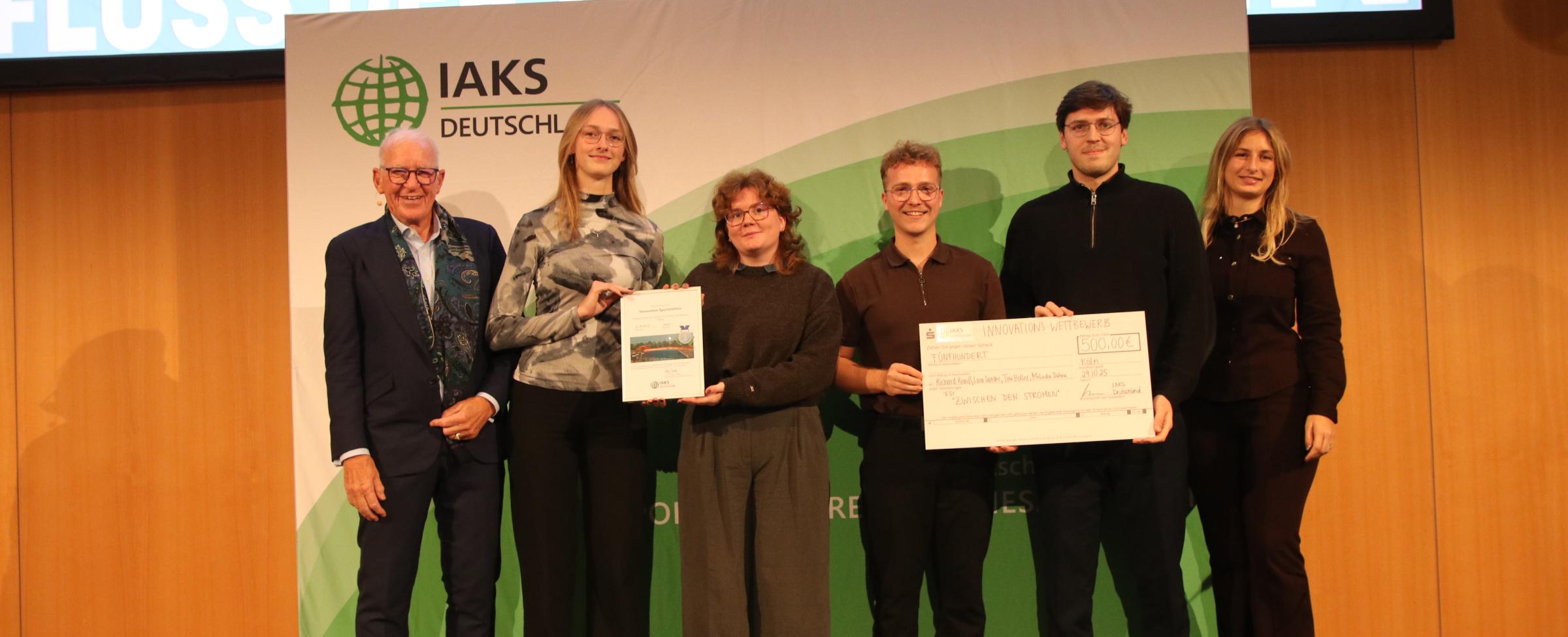 3 Menschen auf Bühne mit Urkunde und Scheck