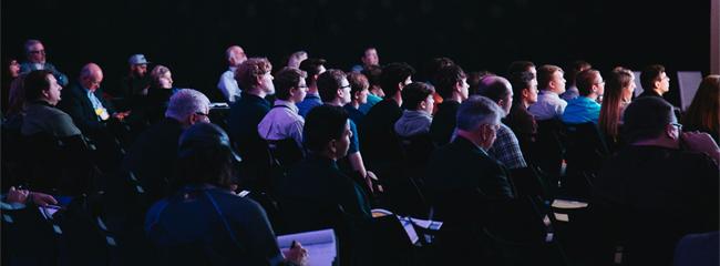 viele Menschen sitzen in einem dunklen Raum, sind auf eine Bühne ausgerichtet