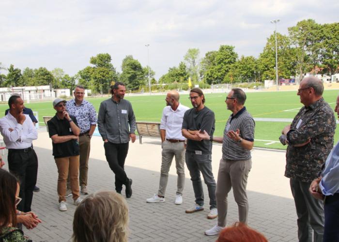 Männer mittleren Alters stehen im Kreis am Rande eines Fußballplatz und diskutieren freundlich