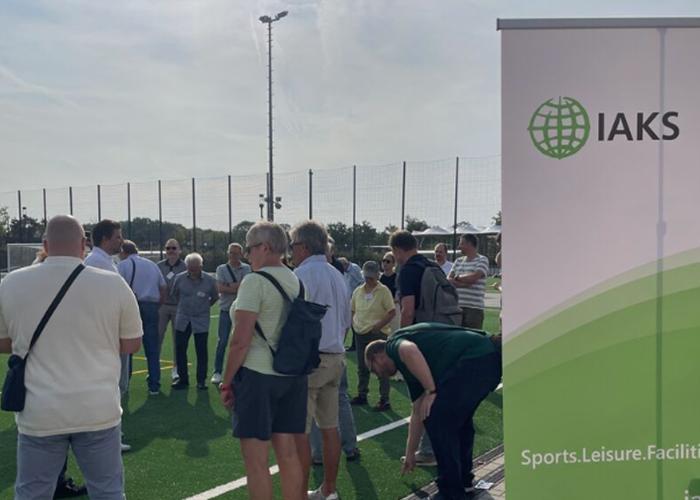 Tagungs-Gäste stehen auf Fussballplatz, in rechter Bildhälfte IAKS Roll Up