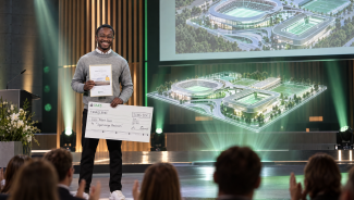 Mann steht auf Bühne Siegerurkunde neben ihm schwebt eine Sportstätte als Hologramm